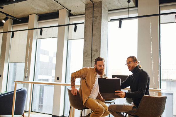 entrepreneur in black turtleneck and eyeglasses showing folder with business project to attentive bearded colleague in modern office with high tech interior, productivity and collaboration concept