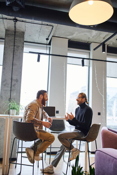 side view of stylish and cheerful business partners discussing startup project while sitting near laptop in contemporary coworking office with large windows, productivity and collaboration concept