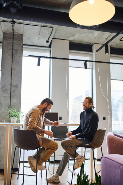 joyful and stylish entrepreneurs planning new business project while sitting near laptop in modern office environment with high tech interior, productivity and collaboration concept