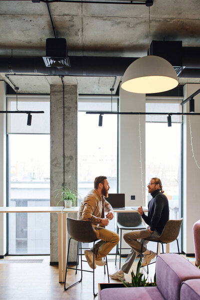 side view of ambitious business colleagues talking about new startup project near laptop in modern office with large windows and high tech interior, productivity and collaboration concept