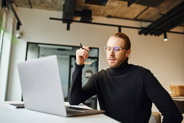 thoughtful entrepreneur in black turtleneck and eyeglasses holding pen and looking at laptop while working on startup planning in contemporary coworking office, business lifestyle concept