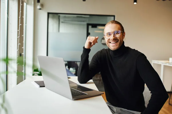 stock image happy entrepreneur in black turtleneck and eyeglasses sitting near laptop on work desk and looking at camera in coworking environment in modern office, business lifestyle concept