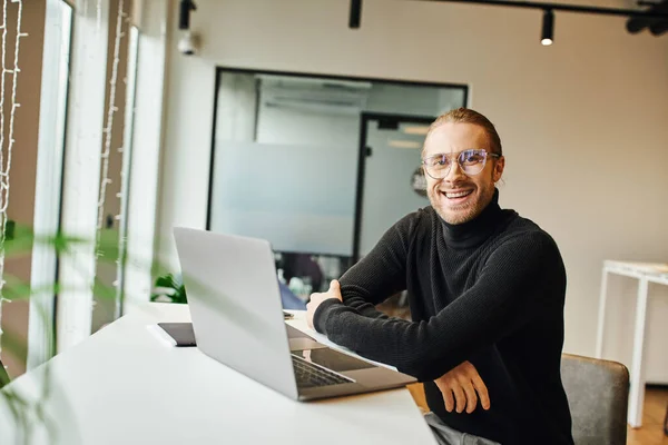 現代的なオフィス環境で折り畳まれた腕でノートパソコンの近くに座っている間にカメラを見て黒のタートルネックと眼鏡で楽しくスタイリッシュなビジネスマン ビジネスライフスタイルの概念 — ストック写真