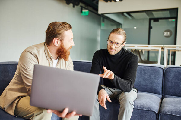 displeased businessman in eyeglasses and black turtleneck pointing at laptop with startup project near bearded colleague while sitting in contemporary office, business partnership concept