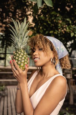 Gülümseyen ve şık Afro-Amerikan kadın portresi başörtüsü takmış elinde taze ananas olan ve bulanık serada dikilirken yüzünü çeviren, tropikal vahadaki moda düşkünü kadın.