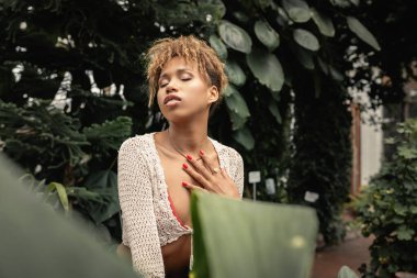 Genç ve şık Afro-Amerikan kadın yazın örgü örüyor göğsüne dokunuyor ve gözlerini kapatıyor arka planda kapalı bir bahçede dikiliyor, moda tutkunu tropikal flora ile kaynaşıyor.