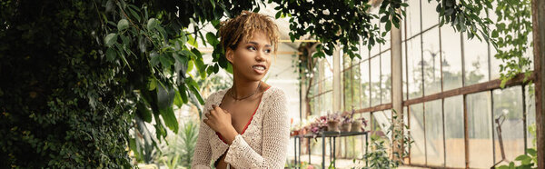 Smiling young african american woman in summer knitted top looking away while standing near green plants in garden center, stylish woman enjoying lush tropical surroundings, banner 