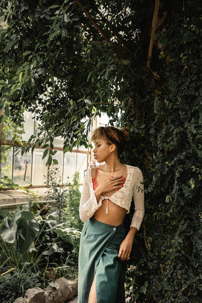 Side view of modern young african american woman in knitted top and skirt touching chest and standing near plants in indoor garden, stylish woman enjoying lush tropical surroundings