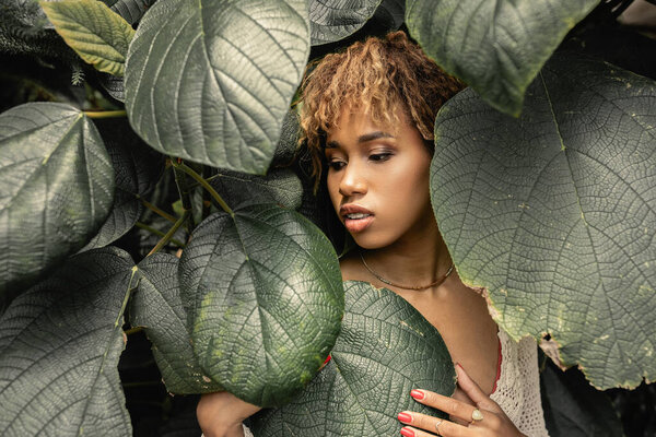 Young and modern african american woman with makeup touching and looking at green foliage while standing in greenhouse, stylish woman enjoying lush tropical surroundings