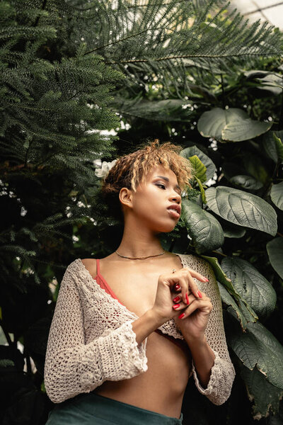 Low angle view of modern young african american woman in summer knitted top looking away while standing near plants in indoor garden, fashion-forward lady in tropical oasis