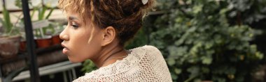 Yazın Afrikalı Amerikalı genç bir kadının arka planda bulanık bahçe merkezinde tropikal bitki örtüsü ve pankartın arasına karışarak vakit geçirirken üstünü örterken yan görünüşü.