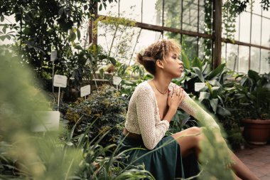Afrika kökenli Amerikalı genç bir kadın şık bir yaz elbisesi içinde arka bahçede yeşil bitkilerin yanında otururken tropikal yeşilliğin ortasında modacı bir kadın, yaz konsepti.