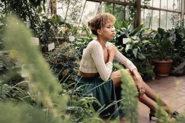 Genç ve rahat bir Afro-Amerikan kadını, yaz kıyafetleri içinde. Arka planda, kapalı alanda yeşil çiçeklerin yanında oturan üstü örülmüş, tropikal yeşilliğin ortasında, modaya uygun bir bayan.