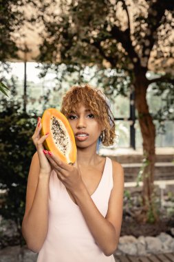 Yazlık papaya elbiseli şık Afro-Amerikan kadın elinde taze papaya ve kameraya bakarken kapalı alanda bulanık duran şık bayan moda ve doğa karışımı, yaz konsepti