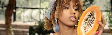 Türbanlı genç Afro-Amerikan kadının portresi taze kesilmiş papaya tutarken arka planda bulanık bir bahçede moda ve doğayı harmanlayan şık bir kadın, afiş 
