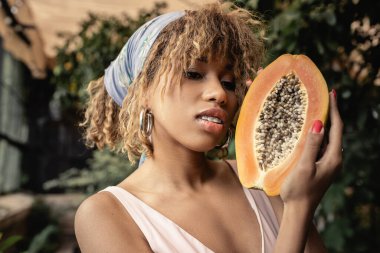 Portrait of trendy young african american woman in headscarf and summer outfit holding cut and ripe papaya while standing in blurred garden center, stylish lady blending fashion and nature clipart