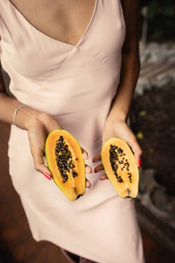 Cropped view of young african american woman in summer dress holding cut and fresh papaya and standing in blurred garden center, fashion-forward lady inspired by tropical plants, summer concept clipart