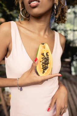 Süslü, genç Afro-Amerikalı kadın. Diş teli takmış, elinde kesilmiş papaya, bulanık bahçe merkezinde. Tropikal bitkilerden esinlenilmiş modacı kadın.
