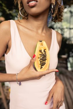 Cropped view of stylish young african american woman with braces wearing summer dress while holding cut ripe papaya in blurred garden center, fashion-forward lady inspired by tropical plants clipart