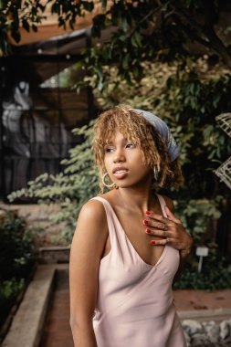 Süslü, Afro-Amerikan kadın portresi başörtüsü ve yaz elbisesi ile göğsüne dokunan, kapalı alanda bulanık duran, yaz kıyafetleri içinde poz veren, yemyeşil tropikal bitkilerin yanında duran kadın.
