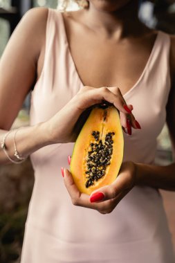 Cropped view of blurred young african american woman in summer dress holding cut papaya while standing in blurred orangery, woman in summer outfit posing near lush tropical plants, summer concept clipart