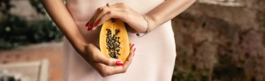 Cropped view of african american woman in stylish summer dress holding cut and juicy papaya while standing in garden center, woman in summer outfit posing near lush tropical plants, banner  clipart
