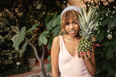 Yazlık diş teli takan, sulu ananas tutan ve bulanık turuncu kameraya bakan neşeli Afro-Amerikalı kadın yaz kıyafetleri içinde, yemyeşil tropikal bitkilerin yanında poz veren kadın.