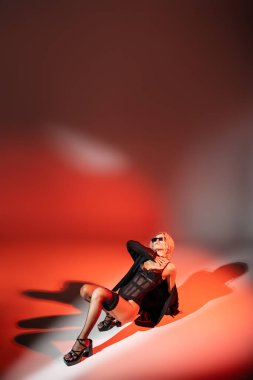 Cinsellik ve moda. Koyu güneş gözlüklü, siyah elbiseli, ceketli, çoraplı, kırmızı ve gri arka planda poz veren topuklu sandaletli baştan çıkarıcı bir kadın.