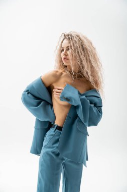 Tahrik edici ve seksi bir kadın. Dalgalı sarı saçlı, gümüş kolye takıyor ve üstsüz vücudunda mavi bir takım elbise giyiyor. Çıplak göğsünü eliyle kapatıyor ve gri arka plana bakıyor.