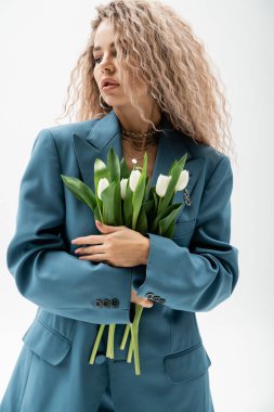 Dişilik ve moda, çekici ve romantik bir kadın. Dalgalı, kül sarısı saçlı, mavi, büyük beden ceketli ve elinde gri arka planda beyaz, taze lalelerden oluşan bir buket var.