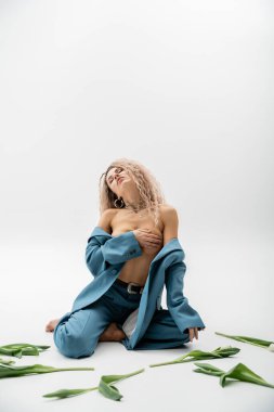 Seksi moda fotoğrafçılığı, boyalı kül sarısı saçlı ve kapalı gözlü tutkulu bir kadın çıplak göğsünü elleriyle örterken gri arka plandaki lalelerin yanında mavi bir takım elbiseyle oturuyor.