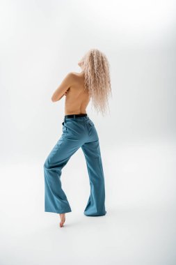 Uzun boylu, seksi, üstsüz ve yalın ayaklı, küllü sarı saçlı, gri arka planda mavi pantolon giyen, ince vücutlu, bireysellik ve kendini ifade eden bir kadın.