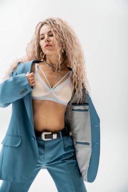 tutkulu sutyen ve mavi büyük beden takım elbiseli kadın gri arka planda duruyor, dalgalı kül sarısı saçlar, moda gümüş aksesuarlar, ince vücut, seksi görünüş, modern moda fotoğrafçılığı.