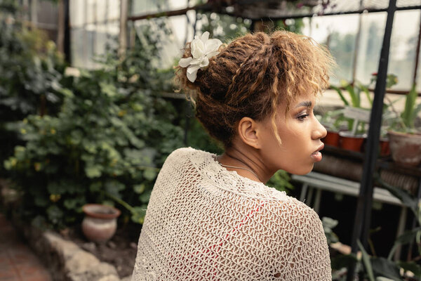 Side view of trendy african american woman in summer knitted top looking away while spending time in blurred indoor garden with green plants at background, fashionista blending in with tropical flora