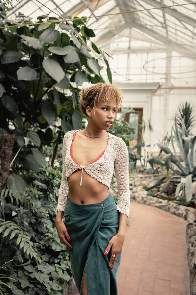 Fashionable young african american woman in summer outfit and knitted top looking away while standing near green plants in garden center, stylish woman with tropical backdrop, summer concept