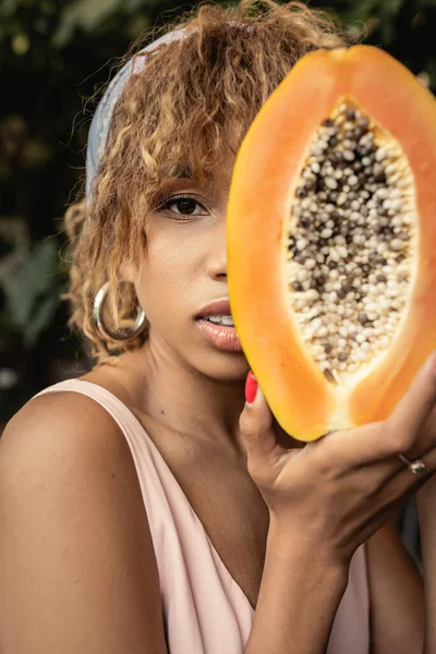 stock image Portrait of young and fashionable african american woman in headscarf and summer dress holding cut and ripe papaya and covering face near plants, stylish lady blending fashion and nature