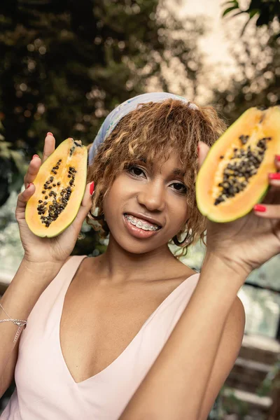 stock image Cheerful young african american woman with braces wearing summer dress and headscarf while holding cut papaya and looking at camera in orangery, fashion-forward lady inspired by tropical plants