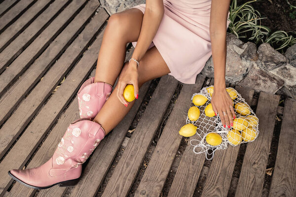 Top view of trendy young african american woman in summer dress and boots holding fresh lemon near mesh bag and sitting in garden center, chic woman in tropical garden, summer concept