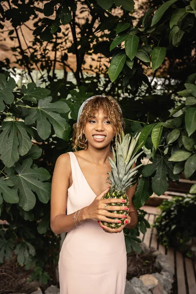 stock image Young and cheerful african american woman with braces wearing summer outfit and holding pineapple while standing near plants in orangery, woman in summer outfit posing near lush tropical plants