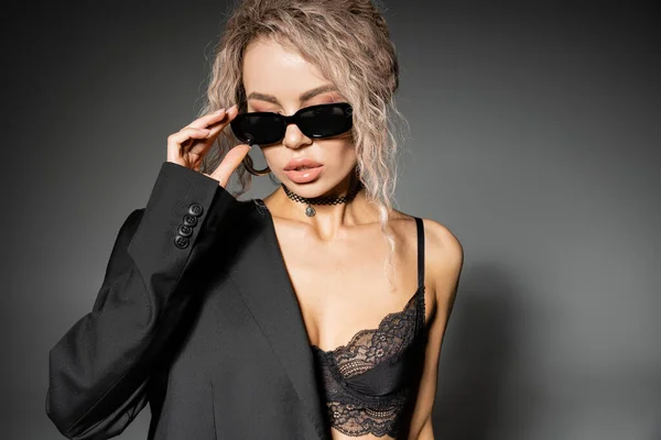 stock image erotic fashion, modern woman with dyed ash blonde hair wearing lace bra and blazer, adjusting dark trendy sunglasses on grey background, glamour style, expressive individuality