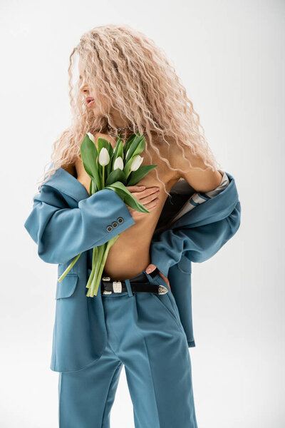 erotic woman with wavy ash blonde hair wearing blue oversize blazer on shirtless body, covering naked breast with hand and holding bouquet of white tulips on grey background, sexy look