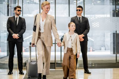 Kişisel güvenlik servisi, resmi giysili ve güneş gözlüklü iki koruma otel girişinin yanında duruyor, mutlu anne ve çocuk el ele tutuşuyor ve bavulla yürüyor, lobiye giriyor, lüks bir yaşam tarzı. 