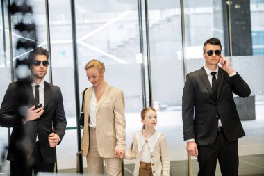 personal security concept, two bodyguards in suits and sunglasses protecting happy mother and child entering lobby, luxury lifestyle, woman and girl holding hands and walking inside  clipart