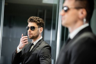 bodyguard service, private security, professionals in suit and sunglasses handsome man standing in hotel lobby and using walkie talkie near work partner, communication, luxury hotel, vigilance  clipart