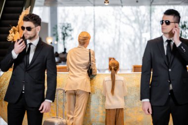 personal security, lifestyle, blonde mother with child standing at reception desk, woman and preteen girl during check in, bodyguards in suit and sunglasses protecting rich family in hotel 