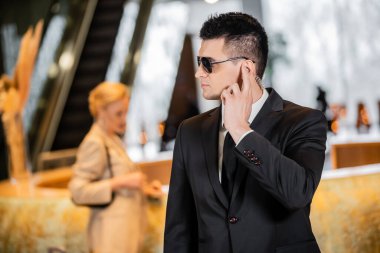 Koruma konsepti, resmi giyimli ve kravatlı yakışıklı bir adam otel lobisinde kulaklığa dokunuyor, güvenlik, iletişim, özel güvenlik, arka plandaki bulanık bayan müşteriyi koruyor. 