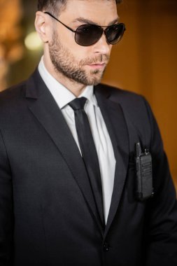 Yakışıklı koruma, kravatlı ve güneş gözlüklü güvenlik görevlisi otelde duruyor, profesyonel vesikalıklar, ceket cebine bağlı radyo vericisi, sakallı adam. 