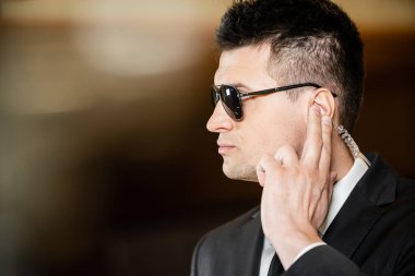 Güneş gözlüklü yakışıklı koruma, takım elbiseli yakışıklı adam otelin lobisinde kulaklığa dokunuyor güvenlik, kariyer, iletişim, tetikte olma, özel güvenlik, otel işi.