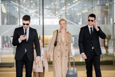 personal security concept, two bodyguards in suits and sunglasses protecting blonde woman with child with luggage entering hotel, wearing autumnal clothes, luxury and rich lifestyle 