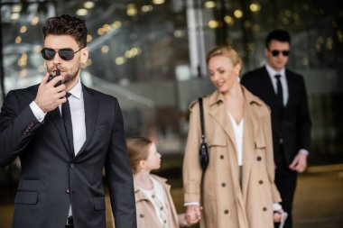 handsome bodyguard communicating through walkie talkie, standing next to successful woman and preteen kid, private security, blonde mother and daughter in trench coats, safety and protection  clipart